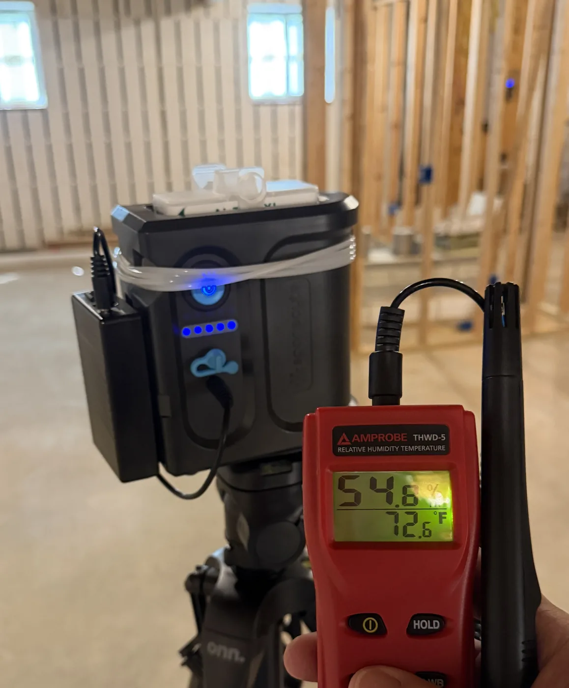 Air sampling pump on a tripod beside a handheld meter reading 54.6 percent relative humidity inside a framed building